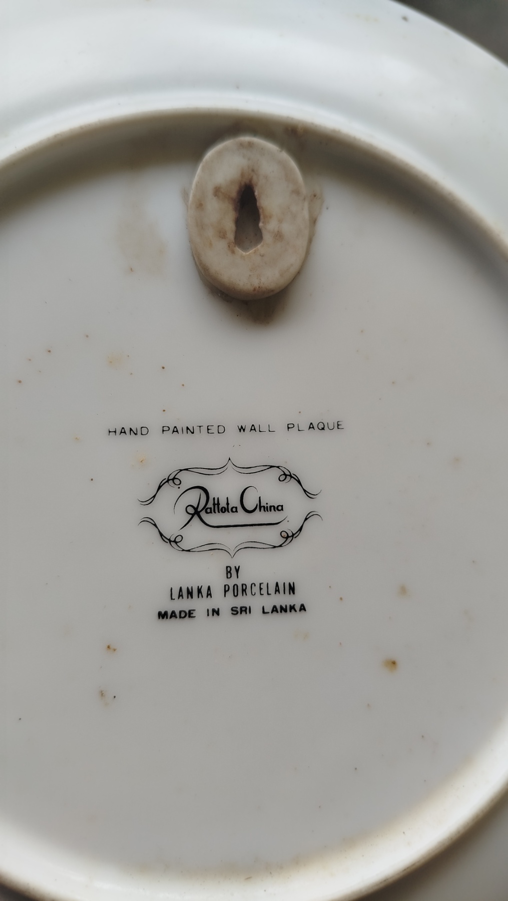 Back view of the porcelain plaque showing the ceramic hanger and the printed inscription: Hand Painted Wall Plaque — Rattota China by Lanka Porcelain — Made in Sri Lanka.